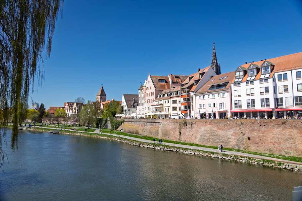 A lush green park along the Danube River in Ulm, Germany, showing the Danube Promenade with people walking, cycling, and sitting on the grass.