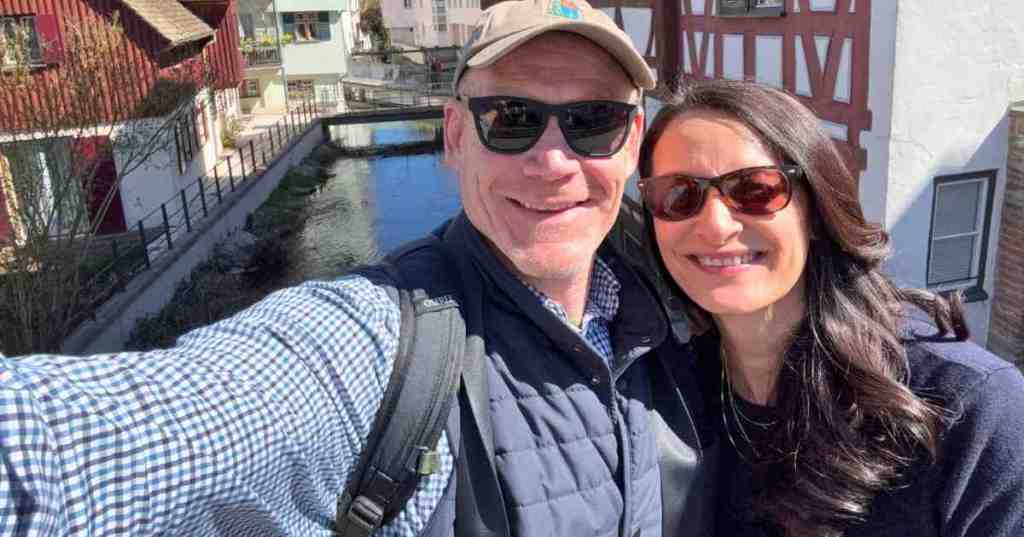A smiling couple taking a selfie on a bridge over the Blau river in the historic Fishermen's Quarter of Ulm, Germany, with traditional half-timbered houses in the background.