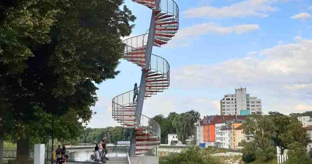 A spiral metal staircase tower known as the Berblinger Turm, leaning over the Danube riverbank in Ulm, Germany, with city buildings and a blue sky in the background.